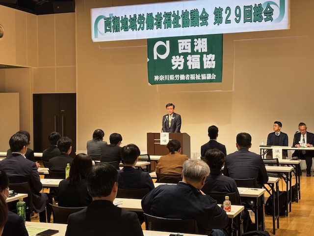 スーツ姿の市長が「西湘地域労働者福祉協議会」と書かれた横断幕の下で演説をしており、多くの人々が机に座ってその話を聞いている総会での写真