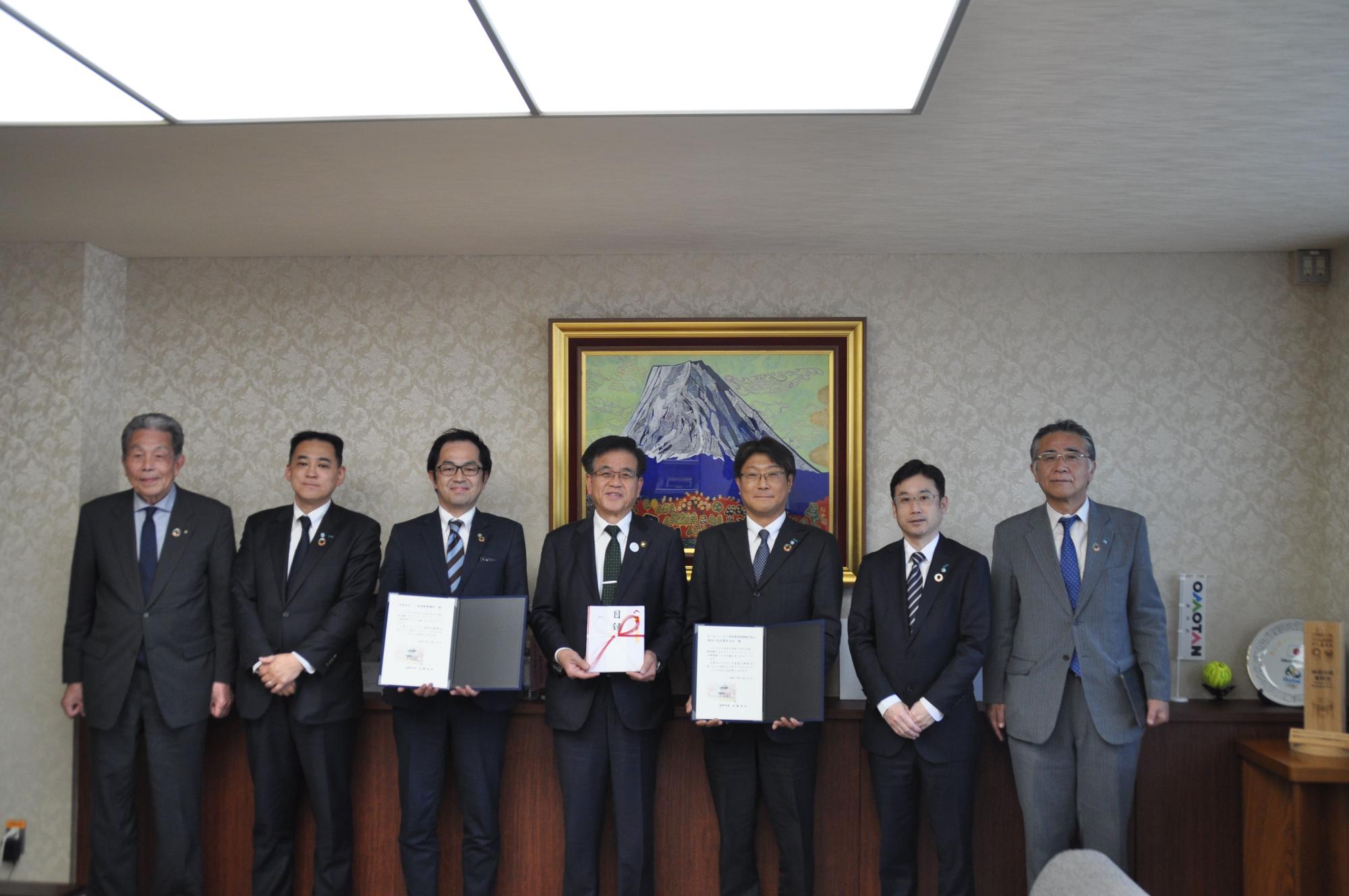 スーツ姿の7人の男性が富士山が描かれた絵画の前で並んでおり、そのうち中央の3人が何か目録や証書のようなものを持っている、記念撮影の様子を捉えた写真