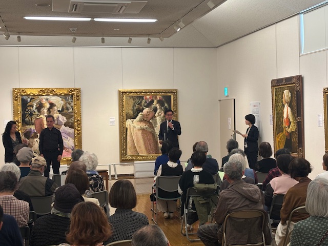 アートギャラリーで、スーツ姿の市長がマイクを手に大勢の聴衆に向かって話しており、背景には複数の絵画が飾られ、別の2人が壇上に立っている、ギャラリートークのようなイベントの様子を捉えた写真