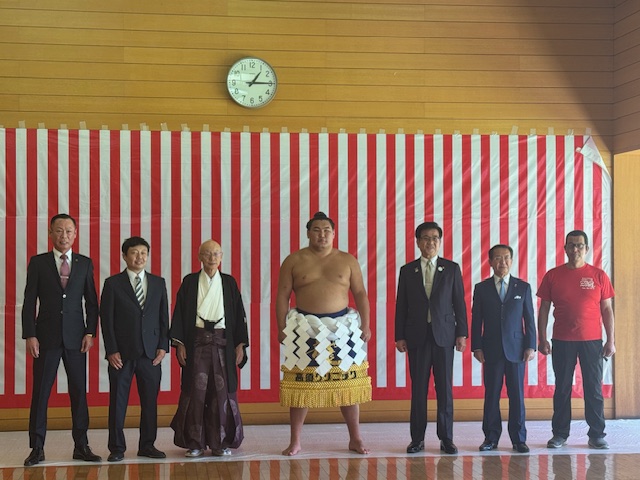 7人の男性が紅白幕の前で並び、その中央にはまわしを締めた力士がおり、両脇にはスーツ姿の男性とTシャツ姿の男性がいる、集合写真を捉えた写真