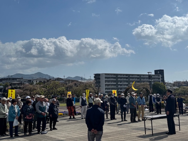 広場で多くの人々が並んでおり、市長がマイクを手に演説をしていて、桜並木や建物、そして大きな月のようなオブジェが背景に見える、屋外で除草作業前の集合をしている様子を写した写真