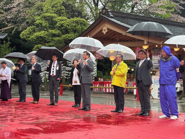濡れた赤いカーペットの上で、傘をさした複数の人々が正面を向いて並んでおり、中央のスーツ姿の市長がマイクを持って話をしている、雨天の神社の境内でイベントが開催されている様子を捉えた写真