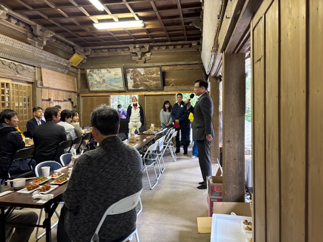 日本の古い木造の建物の中で、スーツ姿の市長がマイクを手にスピーチをしており、テーブルを囲んで座っている多くの人々がその話を聞きながら食事や歓談を楽しんでいる、懇親会のような様子を捉えた写真