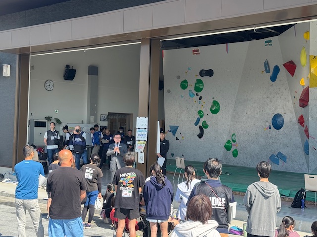 ボルダーハウスのような場所で、スーツ姿の市長がマイクを手にスピーチをしており、その後ろには大勢の観客や競技者らしき人々が真剣な表情でその話を聞いている、ボルダリングイベントの様子を捉えた写真