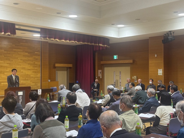 スーツ姿の市長が演台で話をしており、その周りには多くの人々が机と椅子に座って熱心に話を聞いている、室内での会合の様子を捉えた写真