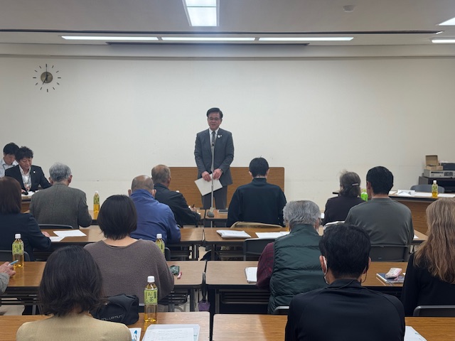 白い壁の会議室で、スーツ姿の市長が演台で話をしており、机に座ってその話を聞いている大勢の聴衆や、壇上に別の関係者がいる、真面目な雰囲気のスポーツ推進委員委嘱式の様子を捉えた写真