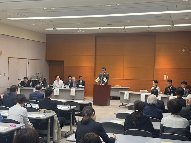 多くの人々が机に向かって座っている部屋で、スーツ姿の市長が演台でマイクを手に話をしており、壇上には数人の男女が並んで着席している、室内での総会の様子を捉えた写真