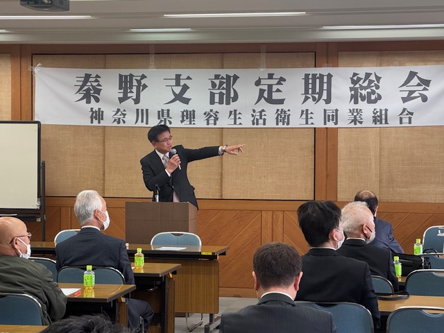 「秦野支部定期総会」と書かれた横断幕の前でスーツ姿の市長がマイクを手に身振り手振りを交えながら話をしており、多くの人々が机と椅子に座り、熱心にその話を聞いている、総会の様子を捉えた写真