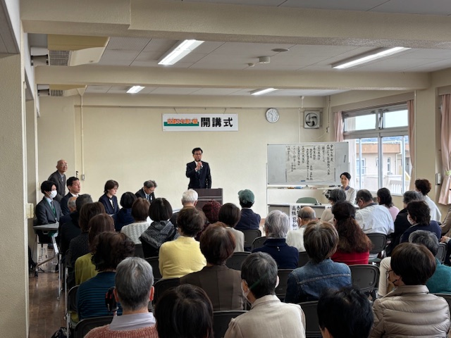広い部屋で、スーツ姿の市長が演台で話をしており、多くの人々が前を向いて着席し、さらに「開講式」と書かれた横断幕が見える、開講式の様子を捉えた写真
