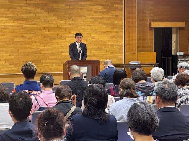 多くの人が椅子に座って前方を見ており、壇上の演台でスーツ姿の市長が聴衆に向かって話をしている、屋内での定期総会の様子を捉えた写真