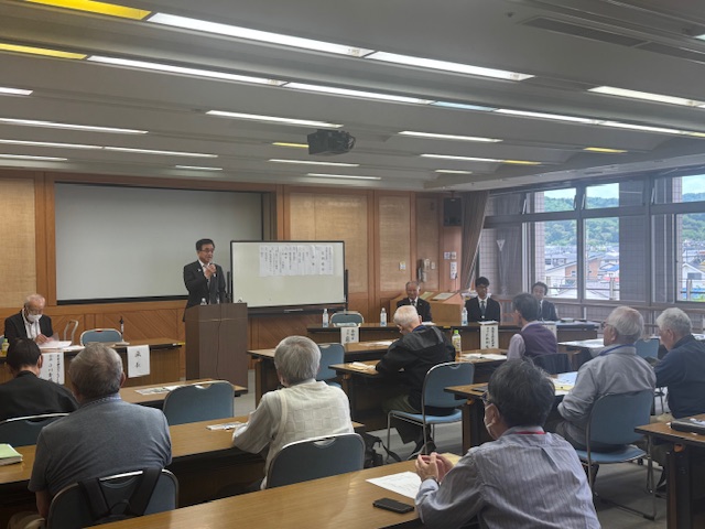 会議室で、スーツ姿の市長が演台で話しており、その周りの机には座って熱心に耳を傾ける人々が多数いる、真剣な雰囲気のボランティアの会総会の様子を捉えた写真