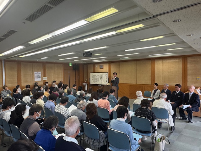 大勢の聴衆が椅子に座って前方を向き、壇上ではスーツ姿の市長が演説をしていて、さらに4人の男女が並んで話を聞いている、室内でのボランティア連絡会総会の様子を捉えた写真