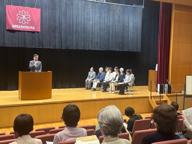 ホールのステージ上でスーツ姿の市長がマイクを手に演説しており、右横には複数の人々が椅子に座って話を聞き、聴衆の人たちが耳を傾けている総会での写真