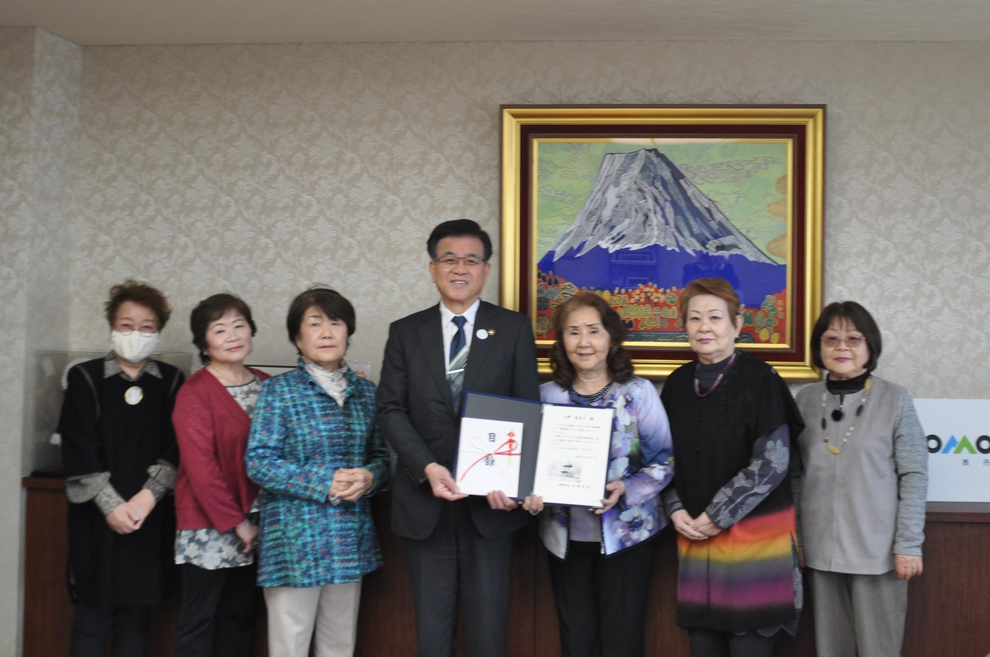 背景に富士山の絵画が飾られた部屋で、スーツ姿の市長が中央に立ち、その両側に6人の女性が並んでおり、男性と右側の女性2人が証書のようなものを持っている、集合写真を捉えた写真