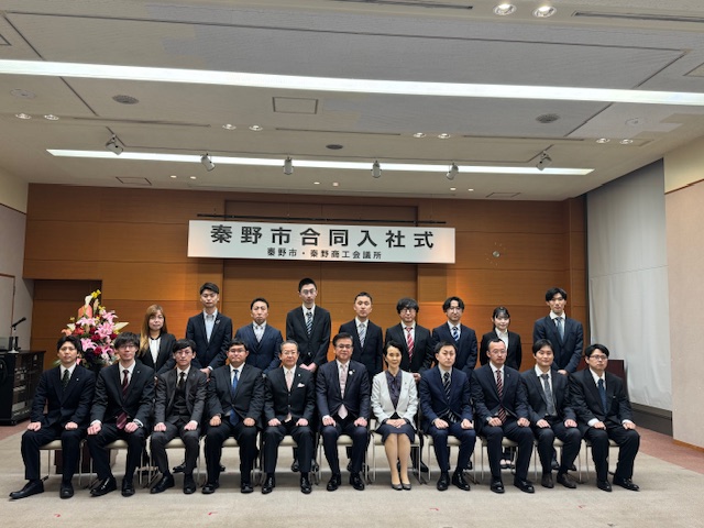 「秦野市合同入社式」と書かれた横断幕の前で、スーツやオフィスカジュアルを着た男性と女性が複数名、椅子に座ったり立ったりしてカメラに向かって並んでいる集合写真を捉えた写真