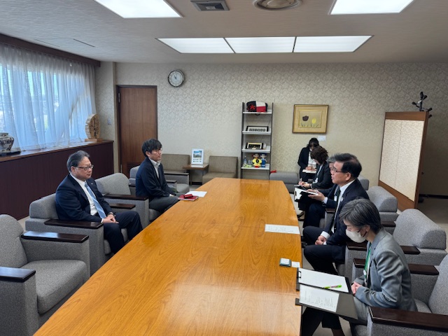 応接室で複数の男女が大きなテーブルを挟んで向かい合って座っており、手前には2人の男性、奥には3人の男女が着席し、真剣な表情で話をしている、意見交換せの様子を捉えた写真