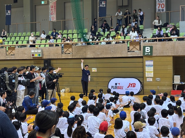 室内スポーツアリーナで、多くの子どもたちが座り、左側にはたくさんの報道陣が集まる中、前に立った紺色のシャツを着た市長が右手を上げて話をしている様子の写真