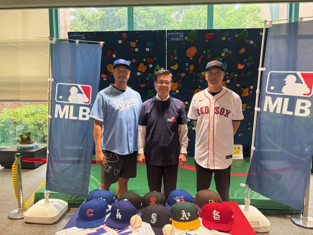 2つのMLBののぼりの間で、野球帽やユニフォームが並べられたテーブルを前に、紺色のシャツを着た市長を中央に、ユニホームを着用したマック鈴木さんと岡島秀樹さんが並んでいる写真