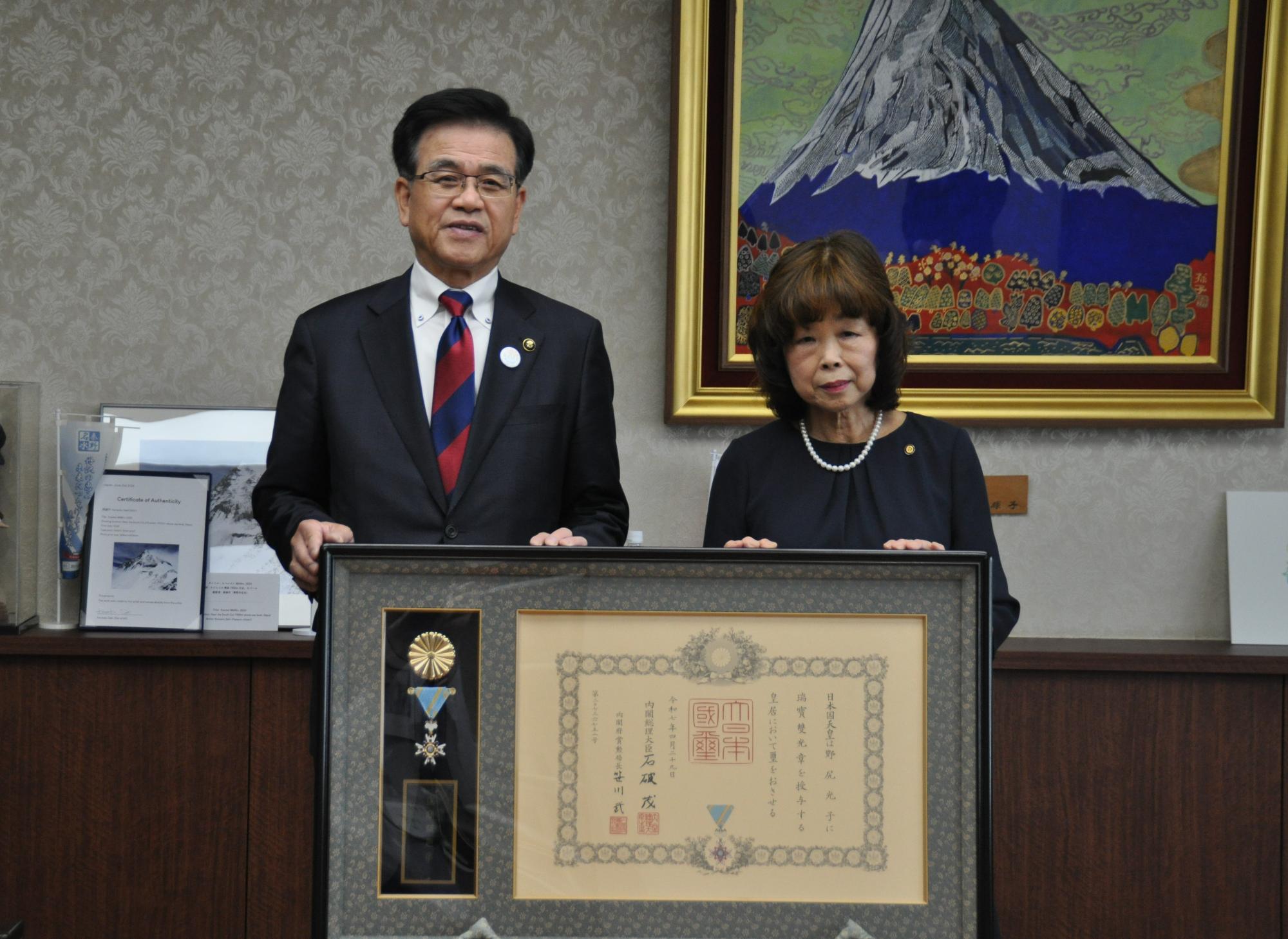 山の絵画が掛かった壁を背景に、スーツ姿の市長と瑞宝双光章を受章した野尻さんが、黒い額縁に入った勲章と表彰状を持ちカメラに向かって並んで立っている記念写真