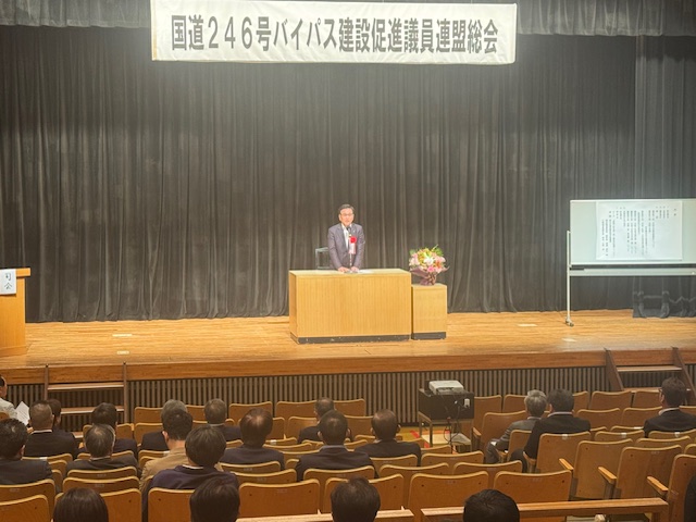 「国道246号バイパス建設促進議員連盟総会」と書かれた横断幕が掲げられたステージの壇上の演台に立つスーツ姿の市長が、客席に座った出席者たちに向かって話をしている写真