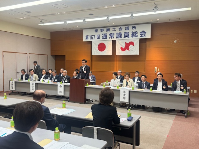 「秦野商工会議所 第157回通常議員総会」と書かれた横断幕と2枚の旗が掲げられた壁の前で、左右の関係者席の中央に置かれた演壇に立った市長が、向かい側に並んだ長机の席に座った出席者に話をしている様子の写真