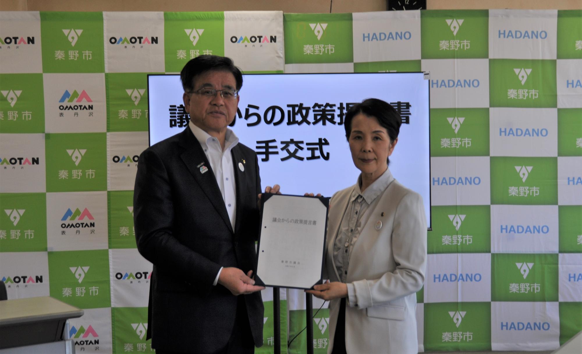 秦野市のバックパネルと「議会からの政策提言書手交式」と表示されたモニターを前に、政策提言書を一緒に持ち、カメラに向かって立つスーツ姿の市長と横山議長の記念写真