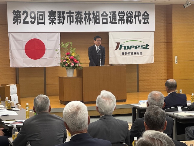 「第29回秦野市森林組合通常総代会」と書かれた横断幕と2枚の旗が掲げられた会場で、席に着いた大勢の出席者を前に、演台に立ったスーツ姿の市長が挨拶をしている写真