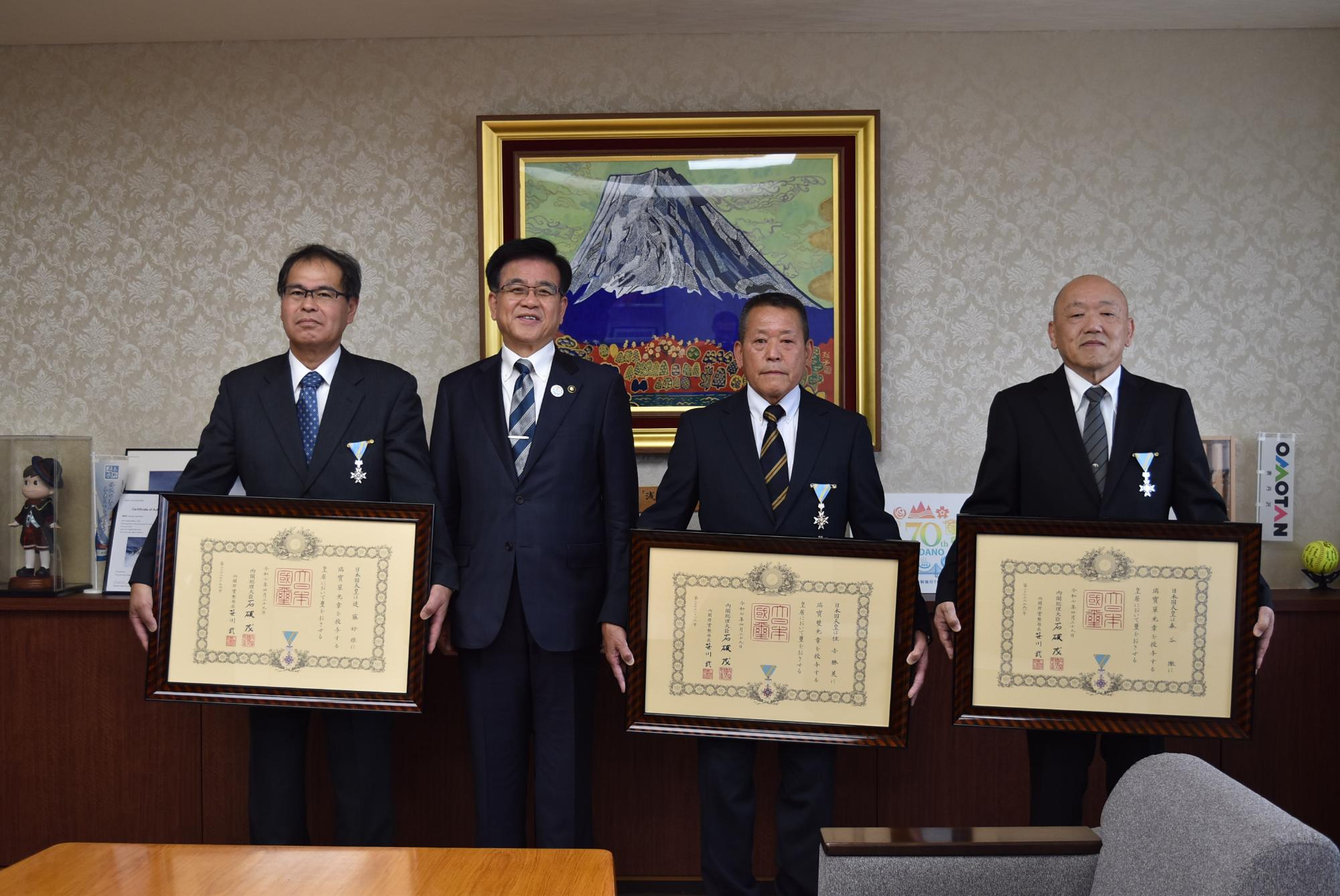 山の絵画が掛かった壁を背景に、市長を挟んで、左胸に勲章をつけた受賞者の男性3名が、それぞれ額縁に入った表彰状を持って並んで立った記念写真
