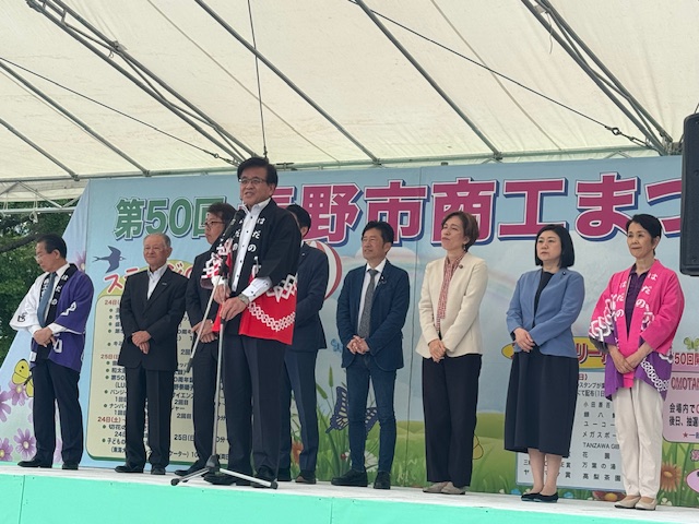 「50回秦野市商工まつり」と書かれたイベント会場の屋外ステージに、8人の関係者の男女が横一列に並んで立ち、その前に置かれたスタンドマイクの前に立った、黒と赤色の法被を着た市長が挨拶を行っている様子の写真