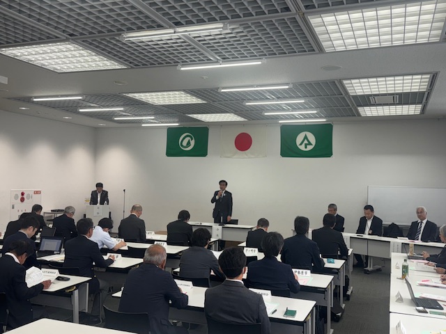 緑のロゴマークの旗2枚と日本の国旗が掲げられた室内で、多くの関係者が長机に向かって座っており、中央の演台に立つスーツ姿の市長がマイクを持って話している会議の様子の写真