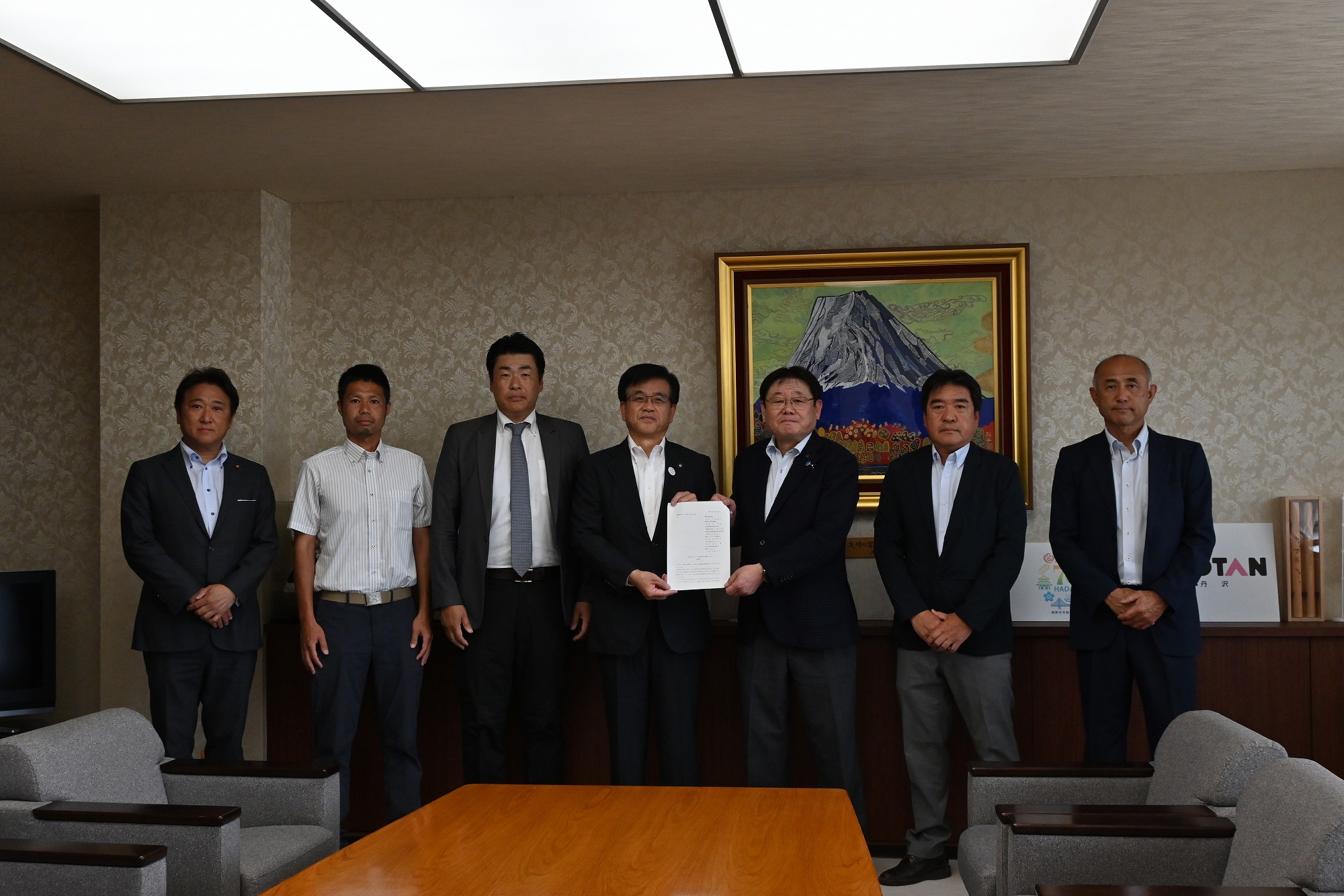 金色の額縁に入った山頂に雪が積もった大きな山の絵画が掛かった壁を背景に、市長を中央に挟んでスーツ姿の6人の男性が並んでおり、市長と代表の男性が一緒に要望書を持っている様子の写真