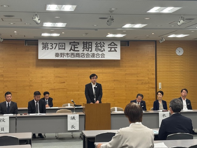 「第37回定期総会 秦野市西商店会連合会」と書かれた横断幕が掲げられた室内で、役員や来賓席の中央に設けられた演台に立った市長が、出席者の方を向いて話をしている様子の写真