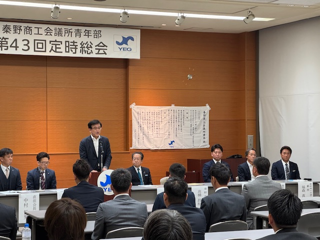 「秦野商工会議所青年部第43回定時総会」と書かれた横断幕が掲げられた会議室で、多くの参加者が席に座る中、関係者席の中央の演台に立った市長がマイクに向かって話をしている様子の写真