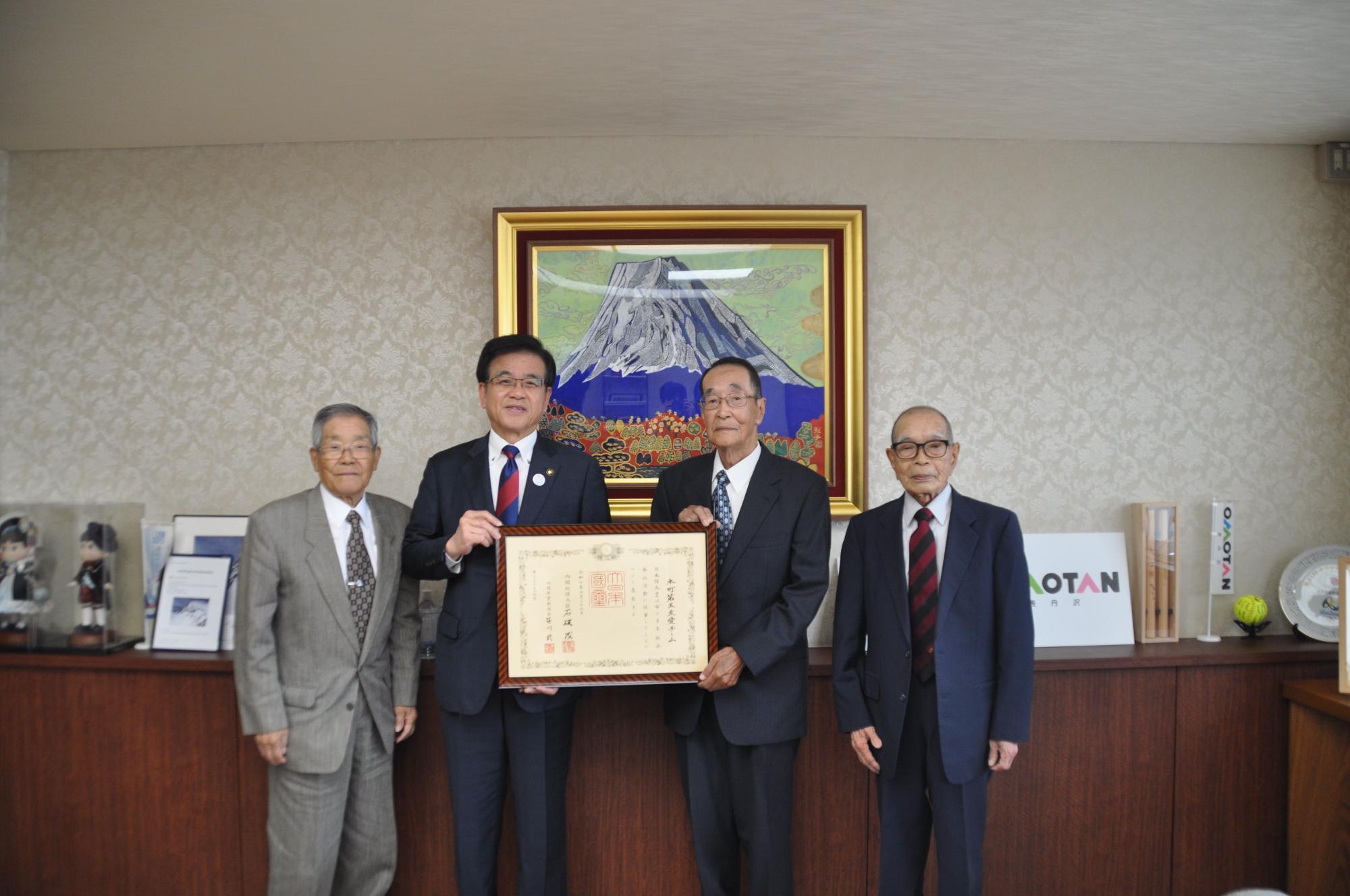 山の絵画が掛かった壁を背景に、4人のスーツ姿の男性が横一列に並んで立ち、中央の市長と代表の男性が一緒に額縁に入った表彰状を持っている記念写真