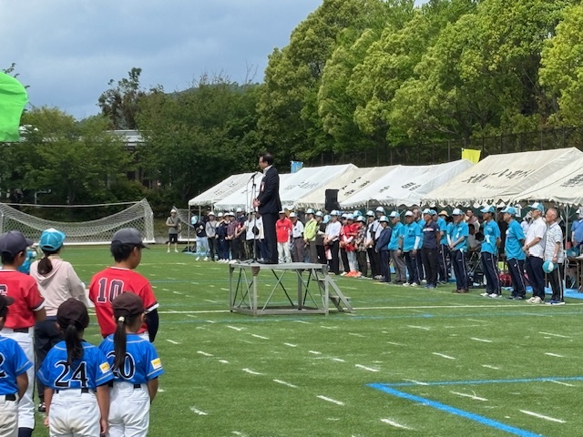 白いテントが並ぶ人工芝のグラウンドで、並んだ白のテントの前に整列した、白と水色のお揃いのキャップを被った関係者などを背に、マイクスタンドが設置された台の上に立つスーツ姿の市長が、前に整列した野球のユニフォームを着た子どもたちの方を向いて話をしている様子の写真