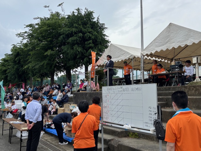 木々や並んだ白いテントを背に、フェンスの前に設置されたスタンドマイクに向かって立った市長が、手書きの得点表が記載されたホワイトボードや長机が置かれた階段下の会場に集まったたくさんの来場者に向かって話をしている様子の写真