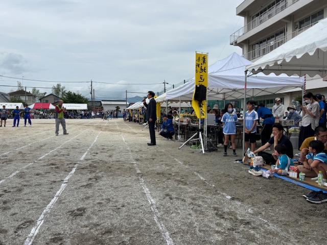 学校の運動場の端に並んだ白いテントを背に、グラウンドに向かってマイクを持って話をしている市長の写真