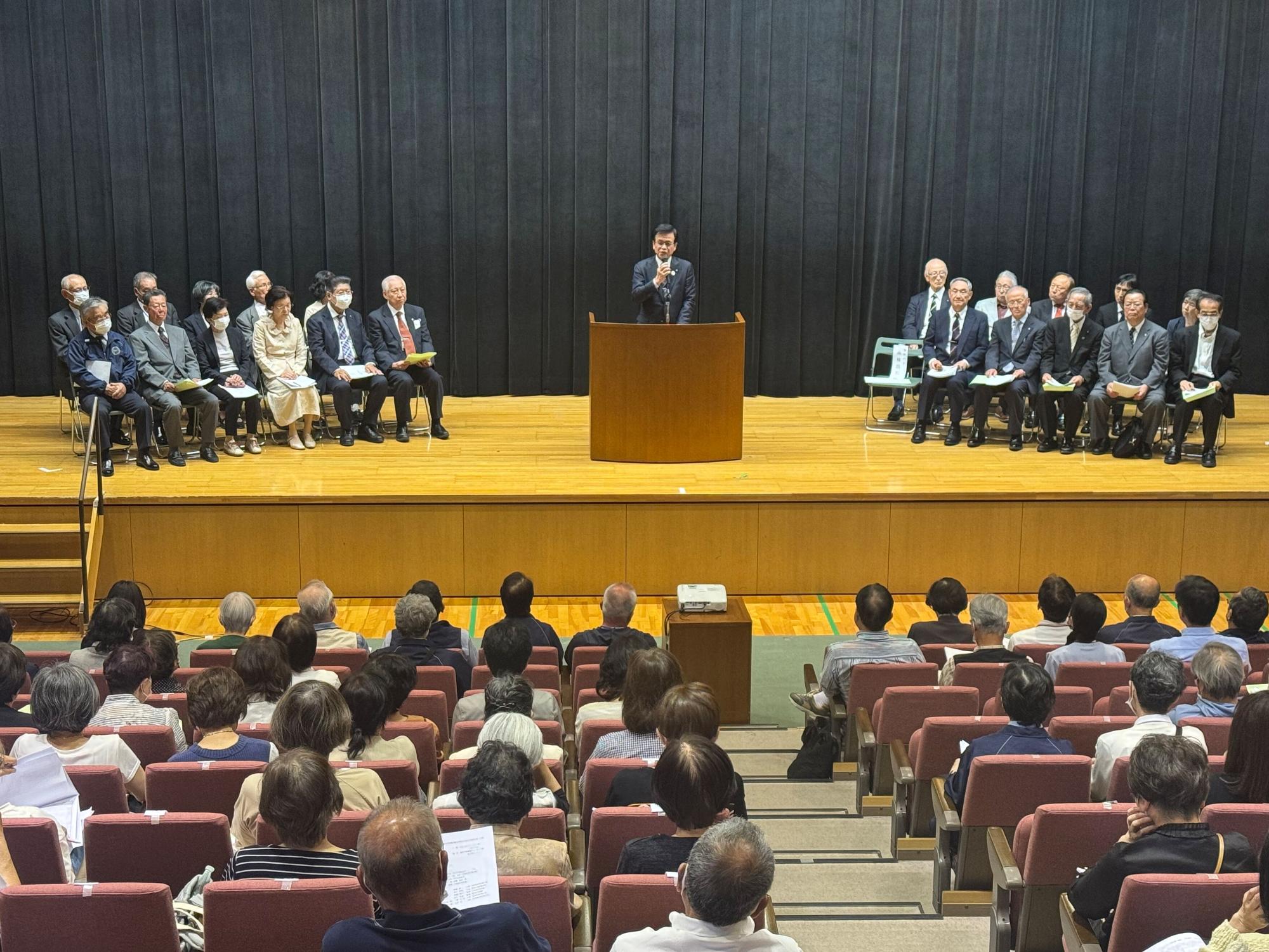 観客席に座っている多くの出席者や、ステージの左右に着席しているたくさんの関係者が、ステージ中央の演台でマイクを持って話をしている市長に注目している様子の写真