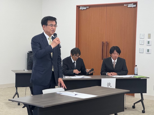 スーツ姿の2人の男性が座った長机の前に置かれた自席から立ち上がったスーツ姿の市長が、マイクを持って話をしている様子の写真