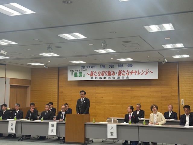 「「進展」～新たな取り組み・新たなチャレンジ～」と書かれた横断幕が掲げられた会場前方の壁を背に配置された、役員や来賓の関係者席の中央にある演台に立った市長が出席者の方を向いて話をしている様子の写真