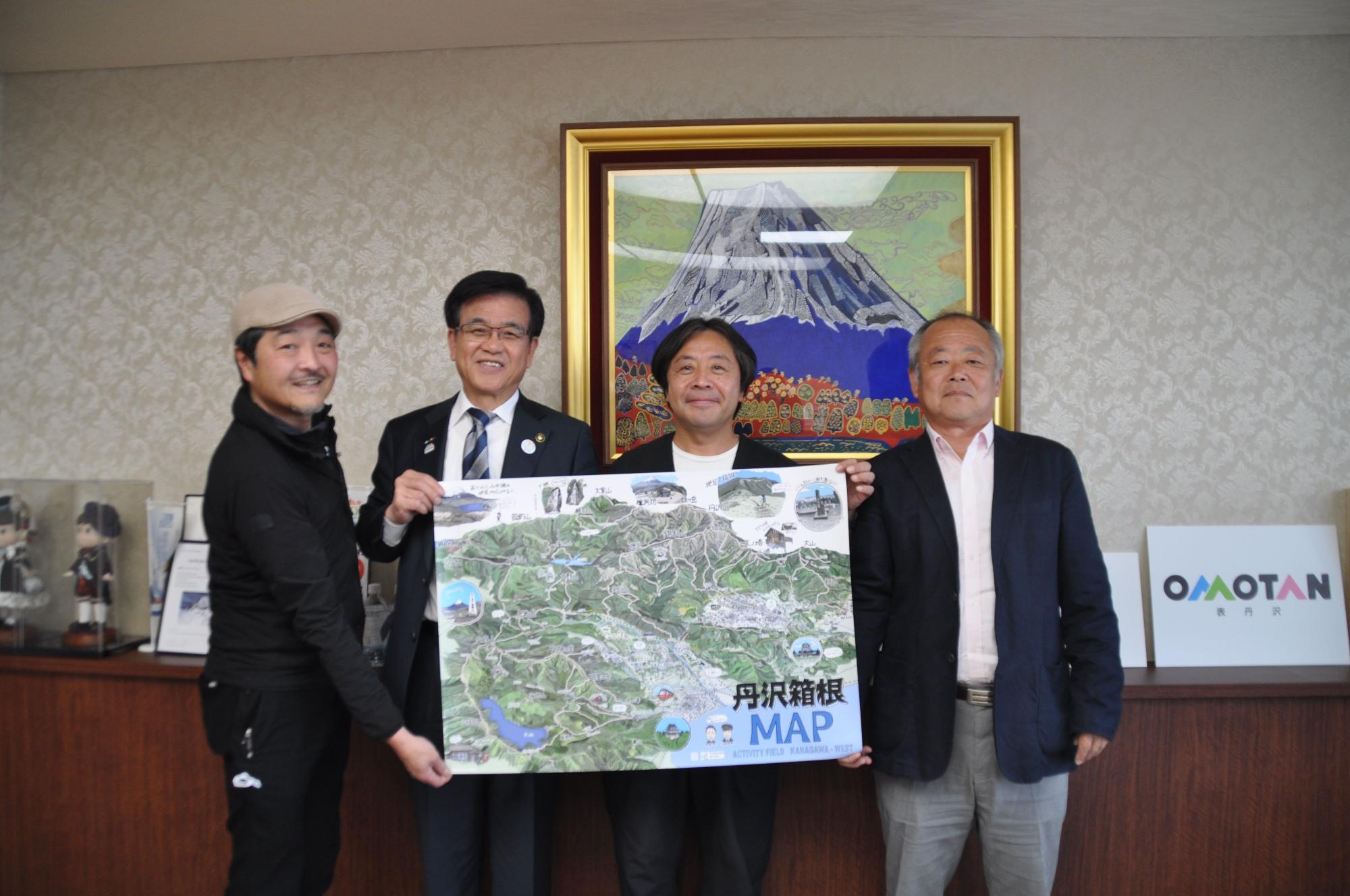 山の絵画が掛かった壁を背景に、4人の男性がカメラに向かって笑顔で並んで立ち、「丹沢箱根MAP」と書かれた地図を中央の市長と2人の男性で持っている記念写真