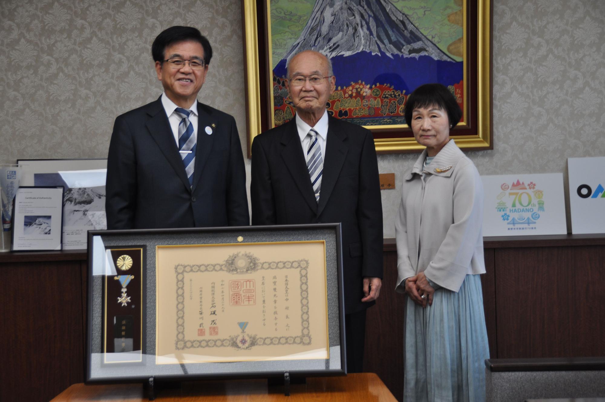 山の絵画が掛かった壁を背景に、市長と受賞者の中村良之さん夫婦が、額縁に入った表彰状と勲章の前に並んで立っている記念写真