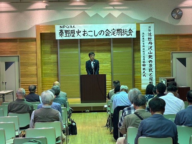 「NPO法人秦野歴史おこしの会定期総会」と手書きで書かれた横断幕が掲げられた小さな舞台上で、演台に立つスーツ姿の市長が多くの出席者を前に挨拶している様子の写真