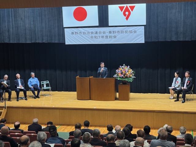 「秦野市自治会連合会・秦野市防犯協会令和7年度総会」と書かれた横断幕が掲げられたステージの左側に3人の関係者が着席し、ステージ下の観客席に座っている大勢の出席者が、壇上の演台に立った市長の挨拶を聞いている様子の写真