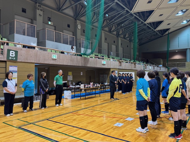 体育館で、関係者が並んで立つ中、中央に立ったスーツ姿の市長がマイクスタンドの前に立ち、向いに立ったスポーツウエア姿の女性に話をしている様子の写真