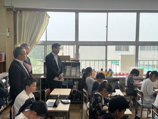 小学校の教室で、生徒たちがタブレットを机に置いて授業を受けている、高学年の授業風景を、教室の後ろに並んで立ったスーツ姿の男性3人が視察している様子の写真