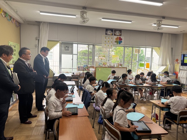 小学校の教室で、コの字型に配置された机でタブレットを使い学習している低学年の生徒たちの授業風景を、教室の後方に並んで立った3人のスーツ姿の男性が視察している様子の写真