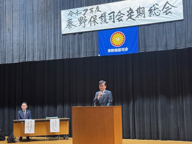 「令和7年度 秦野保護司会定期総会」と手書きで書かれた横断幕が掲げられたステージ左側に関係者の男性が着席しており、演台に立つスーツ姿の市長がマイクを持って挨拶している様子の写真