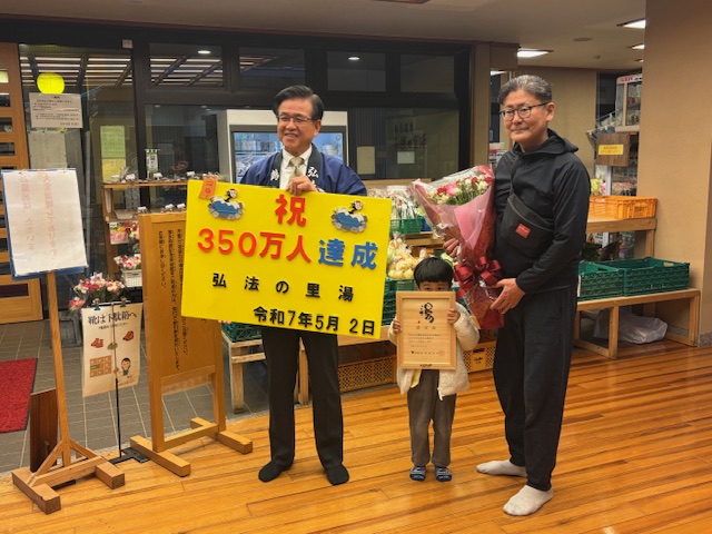 採れたての野菜が入ったかごが並んだ棚の前で、青い法被姿の市長が「祝 350万人達成」と書かれた黄色のパネルを持って立ち、その横に賞状を持った小さな男の子と花束を持った男性が並んで立っている、弘法の里湯の350万人達成を祝う記念写真