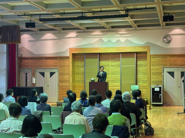 小さな舞台の上の演台で、スーツ姿の市長がマイクを持って挨拶しており、多くの出席者が着席して話を聞いている、レクリエーション協会総会の様子を捉えた写真